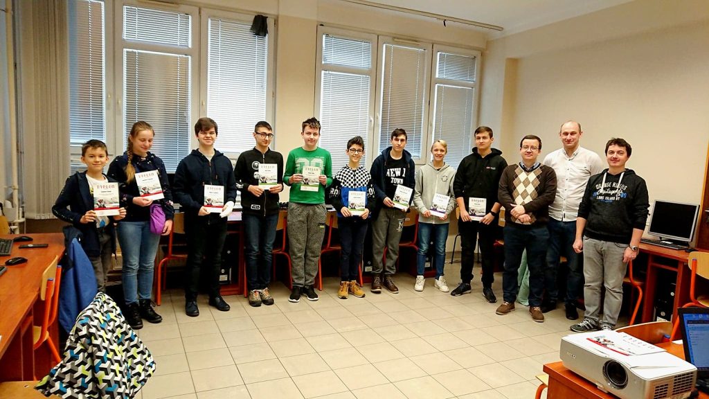 Grupa uczniów i nauczycieli stoi w sali lekcyjnej, trzymając dyplomy. W tle okna i projektor.