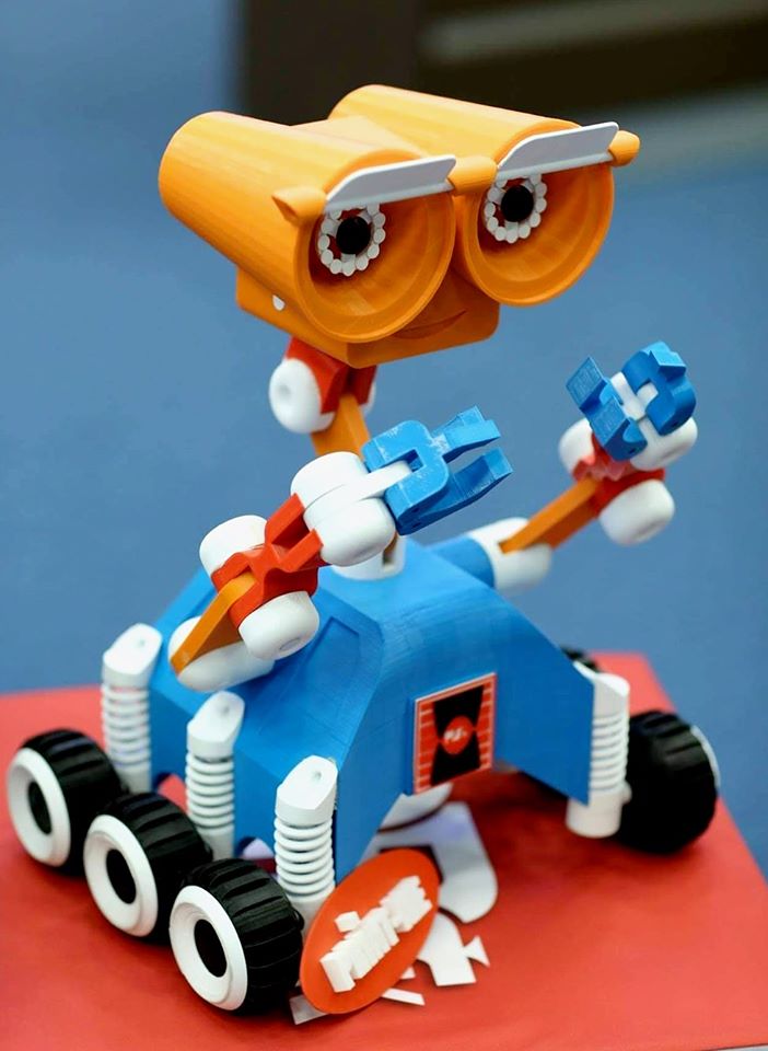 Robot z pomarańczowymi oczami i niebieskimi chwytakami na niebieskiej podstawie z kołami.