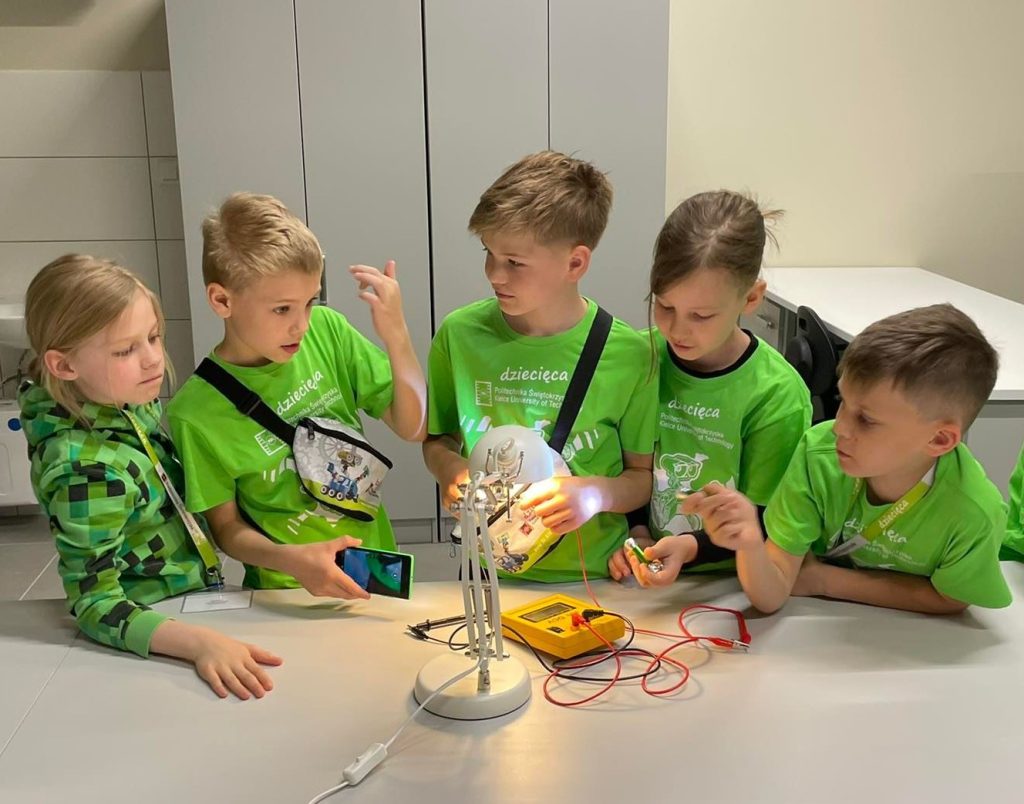 Dzieci w zielonych koszulkach eksperymentują z obwodem elektrycznym. Kluczowe obiekty: dzieci, lampka, multimetr, baterie.