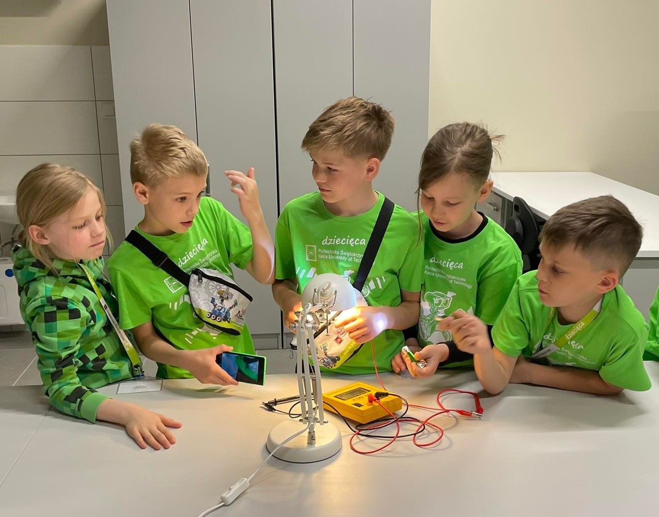 Dzieci w zielonych koszulkach eksperymentują z obwodem elektrycznym. Kluczowe obiekty: dzieci, lampka, multimetr, baterie.