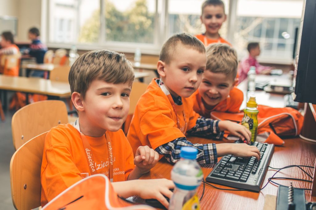 Dzieci w pomarańczowych koszulkach pracują przy komputerach.
